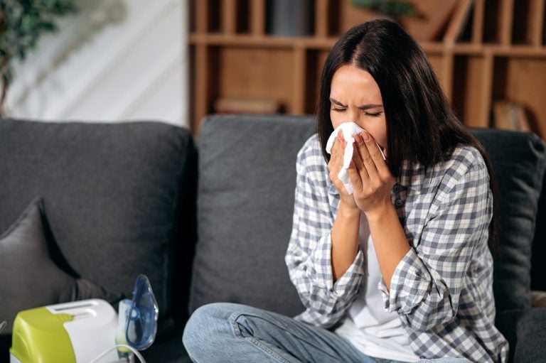 Ill upset caucasian young woman sitting at home on a sofa, blowing running nose, she sneezing on a wipes, sick allergic girl having allergy symptoms, coughing at home, flu, unhealthy